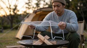 夕暮れのキャンプ場で、日本人女性キャンパーがコールマンのファイヤーディスクにニトリの折りたたみ式キッチンラックを五徳として設置する様子。