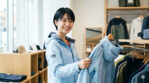 明るいアウトドアショップでヘリーハンセンのジャケットを手に取り笑顔を見せる、若くて可愛いスレンダーな日本人女性のショッピングシーン。