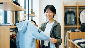 明るいアウトドアショップで、ヘリーハンセンのジャケットを手に取り笑顔を見せる、若くて可愛いスレンダーな日本人女性。