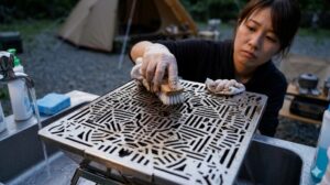おしゃれなデザインの裏に隠れた清掃性の問題。日本人女性が、複雑な幾何学模様の透かし彫りが入った兼用コンロの、隙間に詰まった脂汚れをブラシで落とす様子