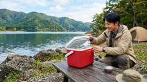 秋の湖畔キャンプ場で、赤いコールマン テイク6クーラーボックスの蓋を開けて中を確認するアジア人男性